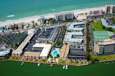Ein Luftbild zeigt eine Strandanlage mit mehreren Gebäuden und einem Pool, umgeben von üppiger Vegetation und einem grünen Kanal.
