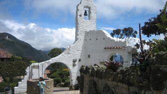 Eine kleine Person steht an einer alten, weißen Mauer mit einer Glocke, umgeben von üppiger Vegetation und Bergen.