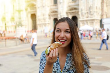 Eine junge Frau genießt einen Cannolo in einer belebten Stadtstraße, umgeben von historischen Gebäuden und Passanten.