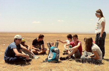 Eine Gruppe von Jugendlichen sitzt im Freien auf trockenem Gras und genießt die Sonne in einer weiten Landschaft.