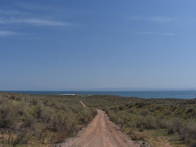 Ein schmaler, staubiger Weg schlängelt sich durch eine grüne Landschaft mit Blick auf einen ruhigen See und blauen Himmel.