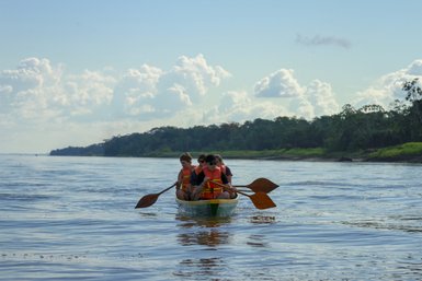 Vier Personen in einem Kanu paddeln auf einem ruhigen Fluss, umgeben von üppiger grüner Vegetation und einem blauen Himmel.