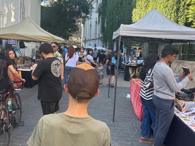 Ein Junge mit einer Kappe steht in einer belebten Straße, umgeben von Marktständen und Menschen, die einkaufen.