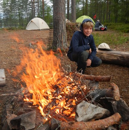 Ein Junge sitzt lächelnd neben einem lodernden Lagerfeuer, umgeben von Bäumen und Zelten in der Natur.