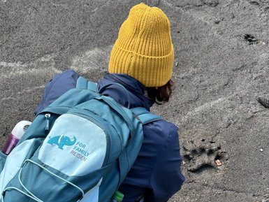Eine Person mit einer gelben Mütze untersucht den Boden, während sie auf einem sandigen Untergrund sitzt.
