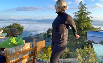 Eine Frau in sportlicher Kleidung steht an einem Glasgeländer und blickt auf eine beeindruckende Berglandschaft mit Wolken.