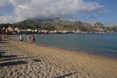 Ein sonniger Strand in Giardini Naxos, wo Menschen am Wasser spazieren und die sanften Wellen genießen.