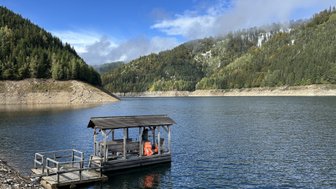 Ein Holzsteg ragt über das ruhige Wasser eines Stausees, umgeben von bewaldeten Hügeln und einem klaren blauen Himmel.
