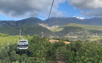 Eine Seilbahn schwebt über üppige grüne Wälder, während majestätische Berge im Hintergrund aufragen.