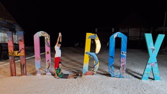 Zwei Kinder posieren fröhlich vor bunten Buchstaben, die das Wort 'Holbox' bilden, auf einem sandigen Strand bei Nacht.