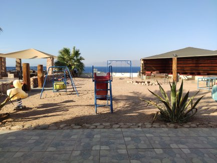 Ein Spielplatz mit blauen Schaukeln und einem gelben Gummienten-Spielgerät, umgeben von Palmen und Meerblick.
