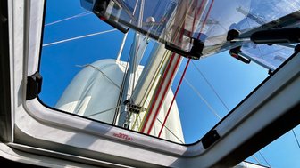 Blick durch das offene Dachfenster eines Bootes, mit Segeln und blauen Himmel im Hintergrund.
