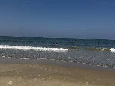 Zwei Personen stehen im flachen Wasser am Strand, umgeben von sanften Wellen und einem klaren blauen Himmel.