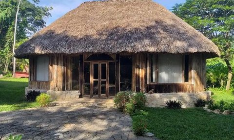 Ein traditionelles, strohgedecktes Haus aus Holz steht in einer grünen Umgebung mit einem gepflasterten Weg davor.