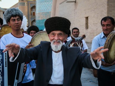 Ein älterer Mann mit einem breiten Lächeln und traditioneller Kleidung tanzt fröhlich in einer Gruppe von Musikern.