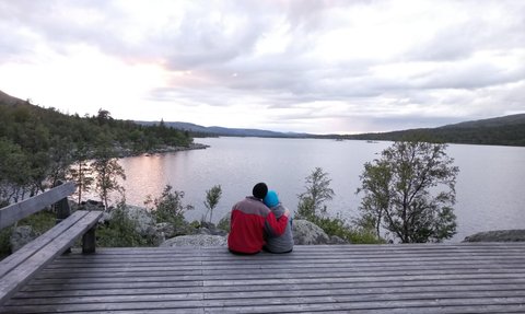 Ein Paar sitzt auf einer Holzterrasse am See und genießt den Sonnenuntergang, umgeben von einer malerischen Landschaft.