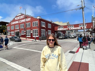 Eine junge Frau steht lächelnd auf der Straße vor dem historischen Monterey Canning Company Gebäude mit roten Wänden.
