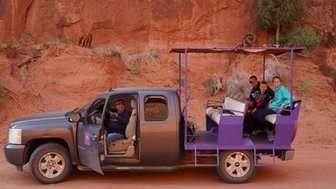 Ein Mann sitzt im Fahrerhaus eines Geländewagens, während eine Familie auf der Ladefläche Platz nimmt, um die Landschaft zu erkunden.