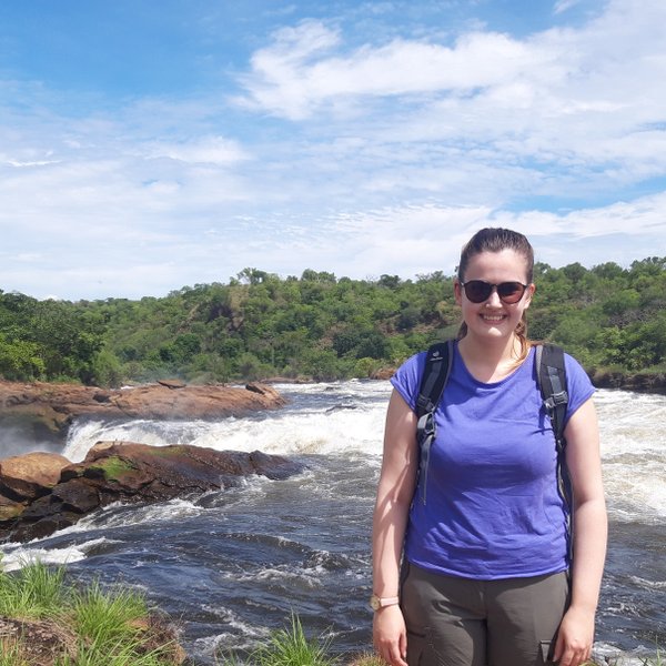 Eine Frau steht lächelnd am Ufer des Murchison Falls, umgeben von üppiger Natur und sprudelndem Wasser.