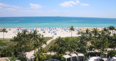 Ein sonniger Strand mit blauen und weißen Sonnenschirmen, umgeben von Palmen und einem klaren, türkisfarbenen Wasser.