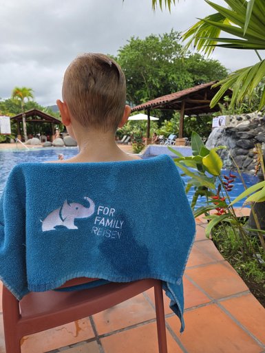 Ein Junge mit nassem Haar sitzt auf einem Stuhl am Pool, ein Handtuch mit einem Elefantenlogo um seine Schultern.