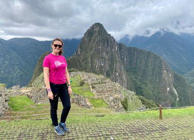 Eine Frau in einem pinken T-Shirt steht vor der beeindruckenden Kulisse von Machu Picchu, umgeben von grünen Bergen.