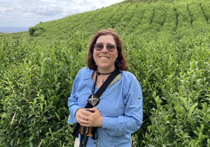 Eine lächelnde Frau steht in einer Teeplantage, umgeben von üppigem grünem Teestrauch und sanften Hügeln im Hintergrund.