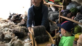 Eine fröhliche Frau und ein Junge arbeiten gemeinsam an einem Wollprojekt in einem hellen, gemütlichen Raum voller Materialien.