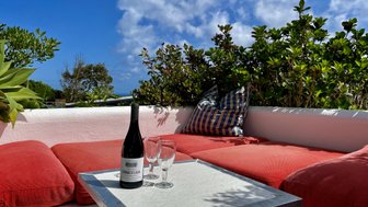 Ein gemütlicher Sitzbereich mit roten Kissen, einem Tisch und einer Flasche Wein, umgeben von üppigem Grün und blauem Himmel.