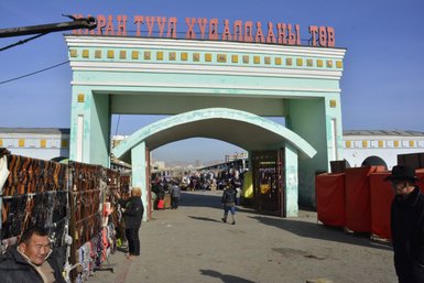 Der Eingang zu einem belebten Markt in Ulaanbaatar, umgeben von Ständen mit verschiedenen Waren und Menschen, die einkaufen.