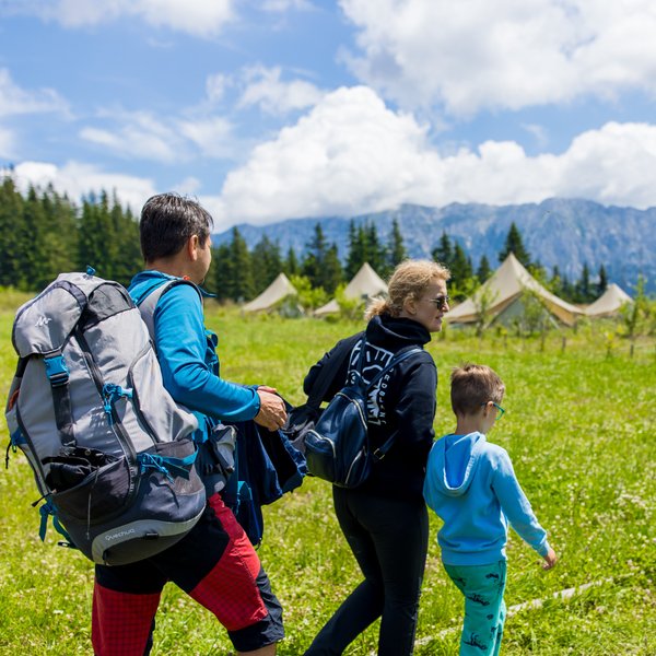 Eine Familie wandert durch eine grüne Wiese, umgeben von Zelten und majestätischen Bergen unter einem blauen Himmel.