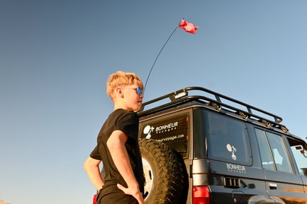 Ein Junge mit Sonnenbrille steht neben einem Geländewagen, während eine Flagge im Wind weht und der Himmel klar ist.