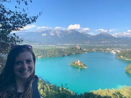 Eine Frau mit langen Haaren lächelt vor einer atemberaubenden Aussicht auf den Bleder See und die umliegenden Berge.