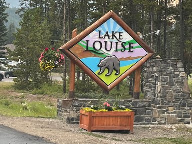 Ein farbenfrohes Schild mit dem Schriftzug 'Lake Louise' und einem Bärenmotiv, umgeben von hohen Bäumen.