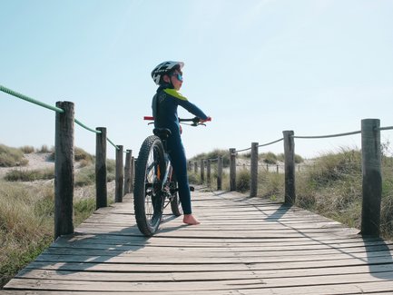 Ein Junge in einem blauen Neoprenanzug steht auf einem Holzsteg und schaut in die Ferne, während er sein Mountainbike hält.
