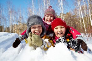 Drei fröhliche Kinder lachen im Schnee, während sie sich in warmen Winterkleidern umarmen und die Sonne genießen.