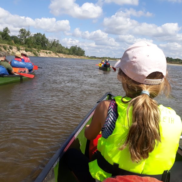 Ein Mädchen mit einem rosa Hut paddelt in einem Kanu, während andere Kanus im Hintergrund auf einem ruhigen Fluss fahren.