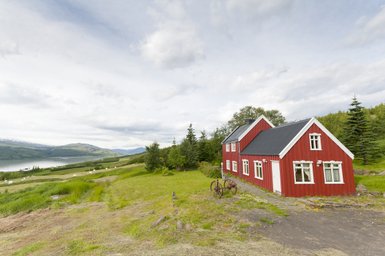 Ein rotes Sommerhaus steht malerisch in einer grünen Landschaft mit Blick auf einen ruhigen See und sanfte Hügel im Hintergrund.