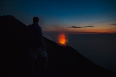 Ein Mensch steht am Rand eines Vulkans und beobachtet die beeindruckende Eruption mit glühenden Lavafontänen im Abendlicht.