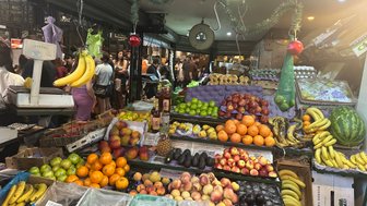Ein lebhafter Marktstand mit einer bunten Auswahl an frischem Obst, darunter Bananen, Äpfel und Orangen, umgeben von Menschen.