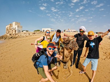 Familie und einheimischer Guide halten einen fröhlichen Moment mit einem Kamel in der Wüste fest – Marokko Familienreise