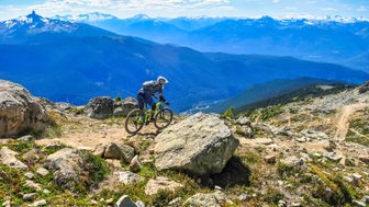 Ein Mountainbiker fährt über einen steinigen Pfad mit beeindruckender Berglandschaft im Hintergrund.