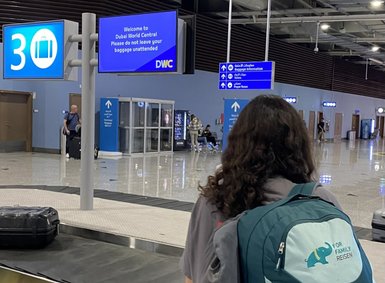 Eine Person mit einem Rucksack steht am Gepäckband in einem modernen Flughafen, während sie auf ihr Gepäck wartet.