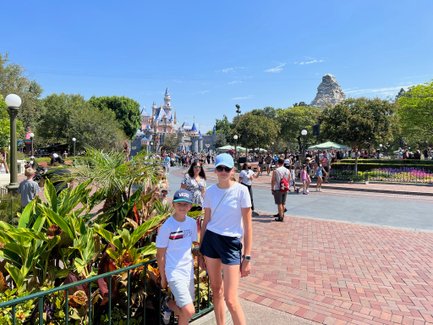 Zwei Kinder stehen vor dem Disneyland-Park, umgeben von bunten Pflanzen und fröhlichen Besuchern an einem sonnigen Tag.