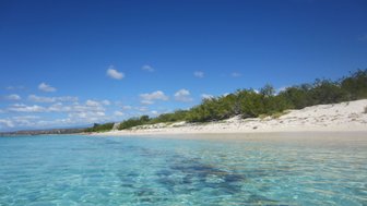 Kristallklares Wasser schimmert in verschiedenen Blautönen, während der weiße Sandstrand sanft in die Küste übergeht.
