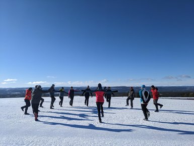 Eine Gruppe von Menschen steht im Schnee und führt eine Aktivität unter blauem Himmel mit wenigen Wolken durch.