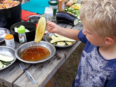 Ein Junge hält eine Zucchinihälfte über eine Schüssel mit Gewürzöl, während er am Tisch mit frischen Zutaten kocht.