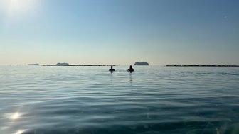 Zwei Personen stehen im ruhigen Wasser, umgeben von einem klaren Himmel und sanften Wellen, während Schiffe in der Ferne sichtbar sind.