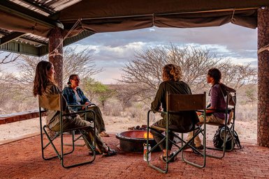 Vier Personen sitzen entspannt um ein Lagerfeuer, umgeben von einer trockenen, offenen Landschaft und einem bewölkten Himmel.