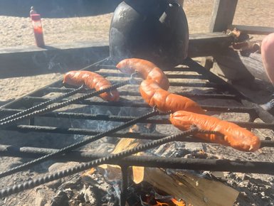 Über dem offenen Feuer brutzeln mehrere Würstchen auf einem Grillrost, während Rauch in die Luft steigt.